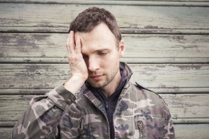Young Caucasian man with headache in camouflage. Outdoor portrait over green rural wooden wall